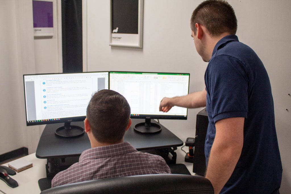 two guys who work in data collaborating over a mailing list. The guy Scott on the left is in his desk chair and the guy Jake on the right is over his shoulder pointing at the computer screen.