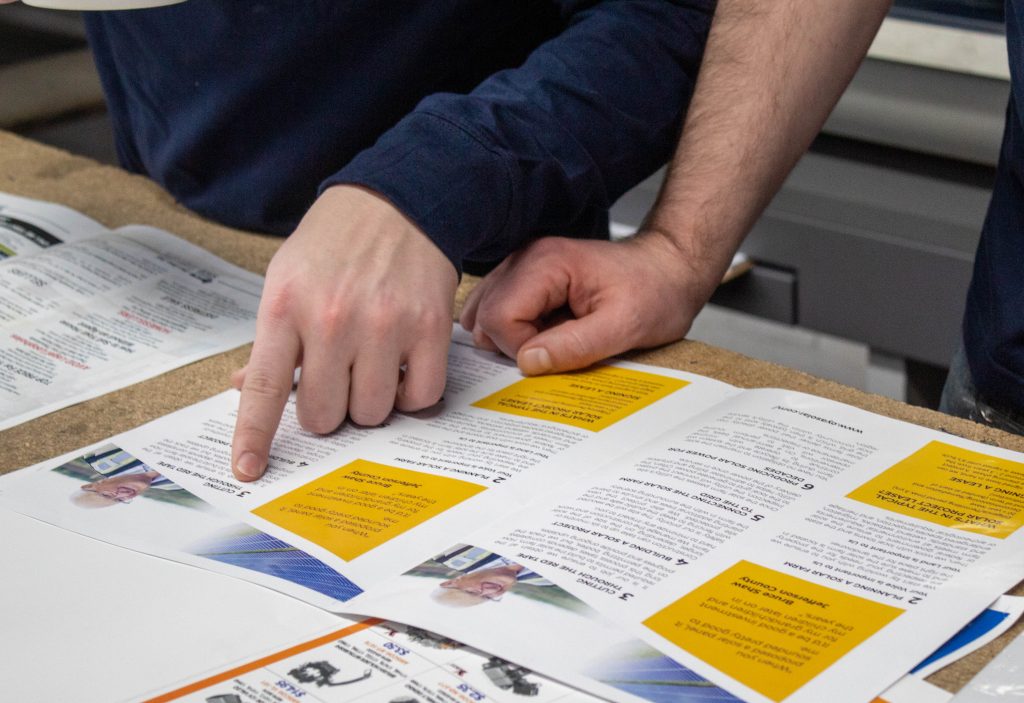 a close up of a printed proof on a table. 2 hands pointing at a certain piece of text on the page.